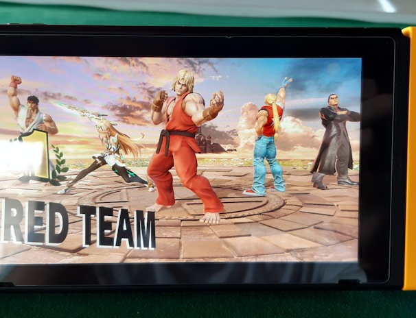 A handheld gaming console displays a screenshot of a fighting game featuring cartoonish characters in dynamic poses. The characters are standing in a virtual arena setting with a sky and cloud backdrop. The console is placed on a green surface.